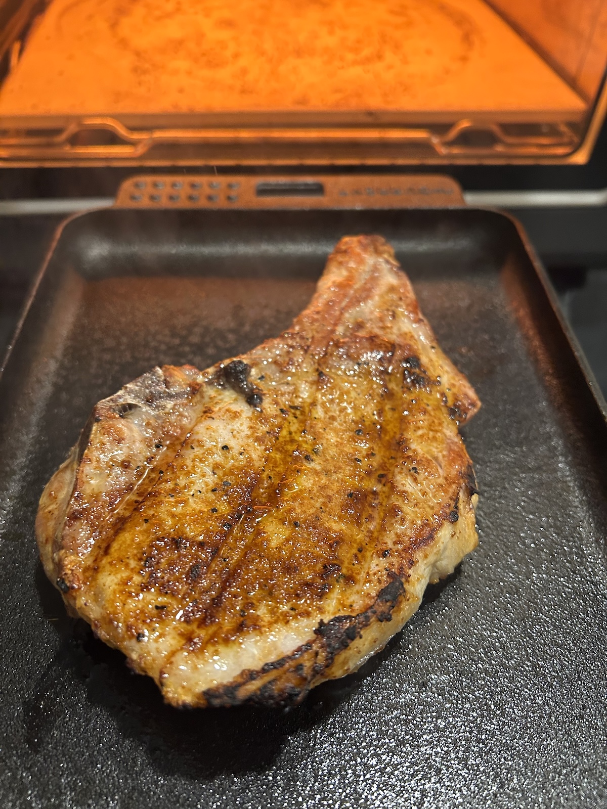 Cooked pork chop shown on a cast-iron sizzle plate in front of Current Model P smart pizza oven