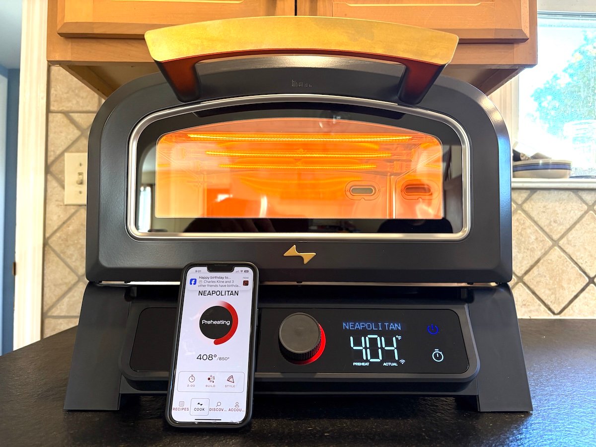 Current Model P smart pizza oven shown from the front with the display reading "Neapolitan 404°F" with a smartphone leaning on the oven front and the phone displaying a similar message on the Current smartphone app.