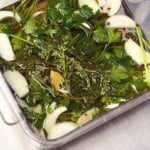 poultry brine in a clear container shown from the top with garlic and herbs visible in the liquid, a wire whisk off to the side.