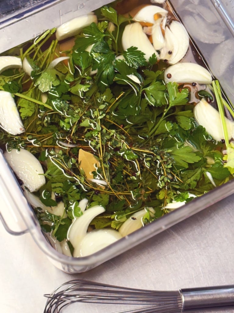 poultry brine in a clear container shown from the top with garlic and herbs visible in the liquid, a wire whisk off to the side.