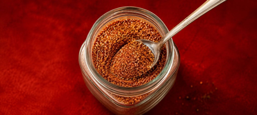 Dry rub seasoning in a glass jar with a spoon inserted.