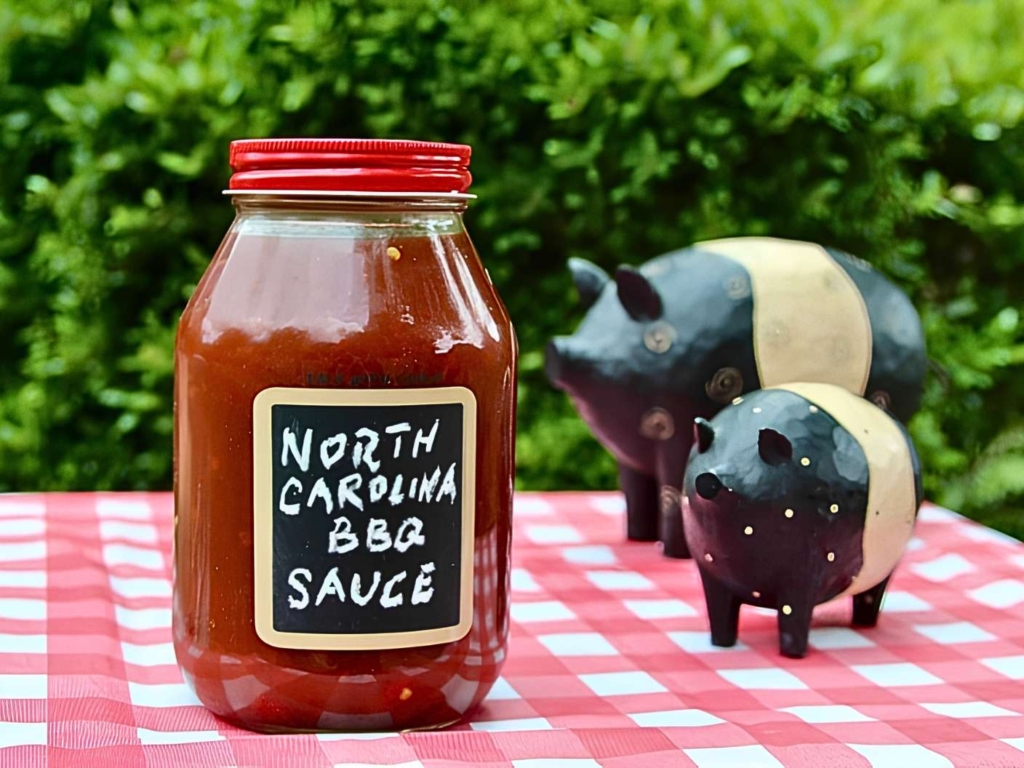 NC Lexington Dip Barbecue Sauce in a labeled glass jar on a chcked tablecloths and two wood pigs
