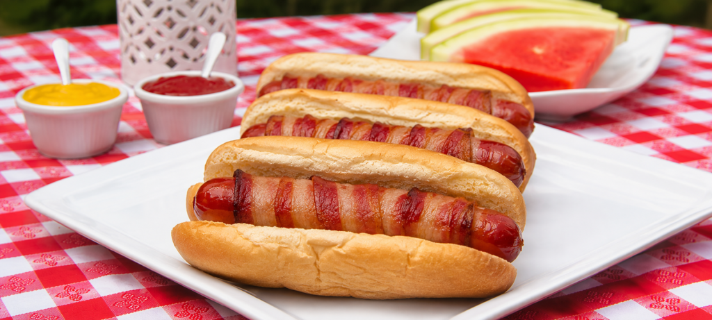 Bacon wrapped hot dogs on a platter with watermelon slices in the background