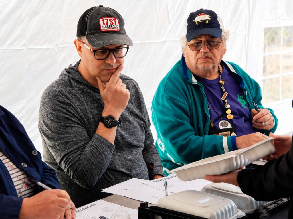 Two men judging meat in a white styrofoam a competition BBQ turn-in box.
