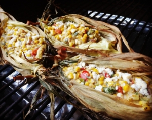elote dip shown in the corn husks on a grill