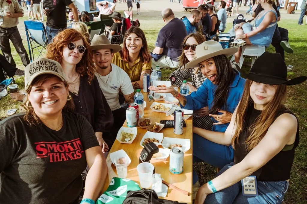 Barbecue festival attendees at the Texas Monthly festival smiling around a picnic table full of barbecue.